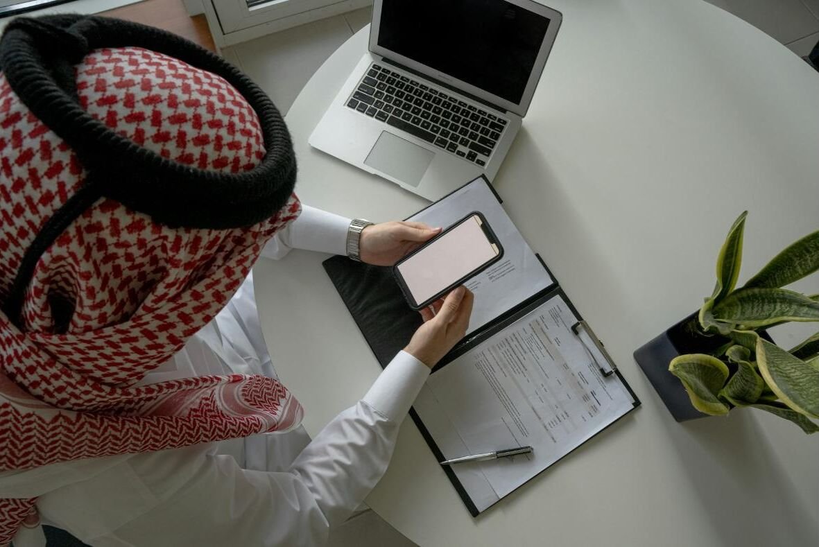 A business owner checking orders on his phone next to a laptop and paperwork
